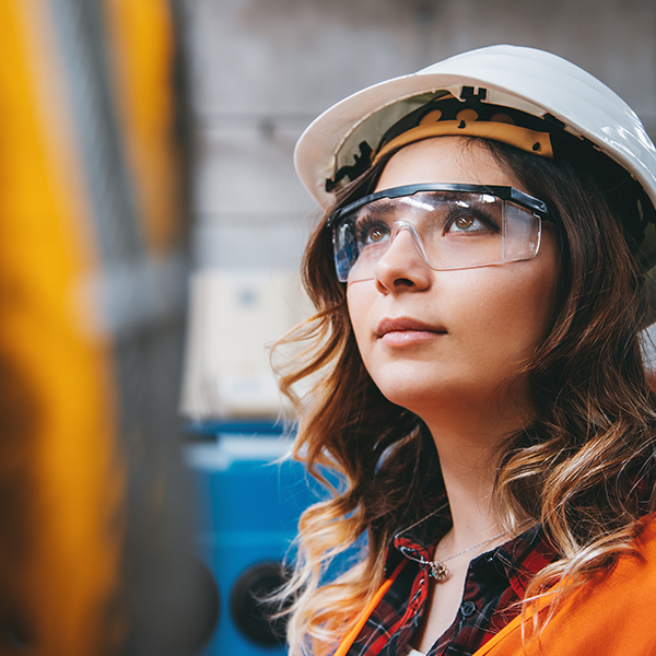 A young female apprentice standing in a factory wearing a hard hat and protective glasses looking up at a machine