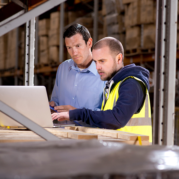 A business man and a man in high visibility clothing stand talking at a computer in a warehouse