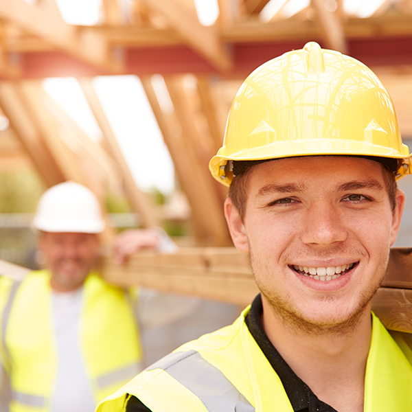 Young male apprentice working on a construction site is carrying wood with a colleague while smiling into the camera